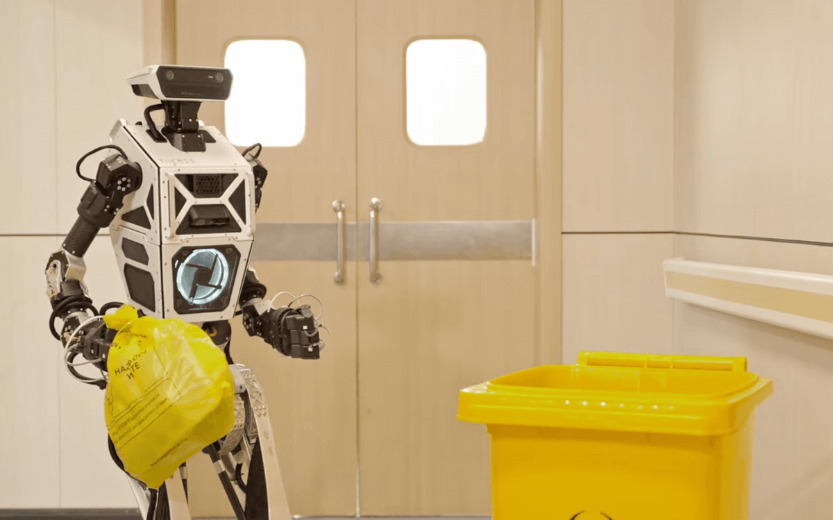 humanoid robot holds small yellow bag near biohazard trash bin in a hallway