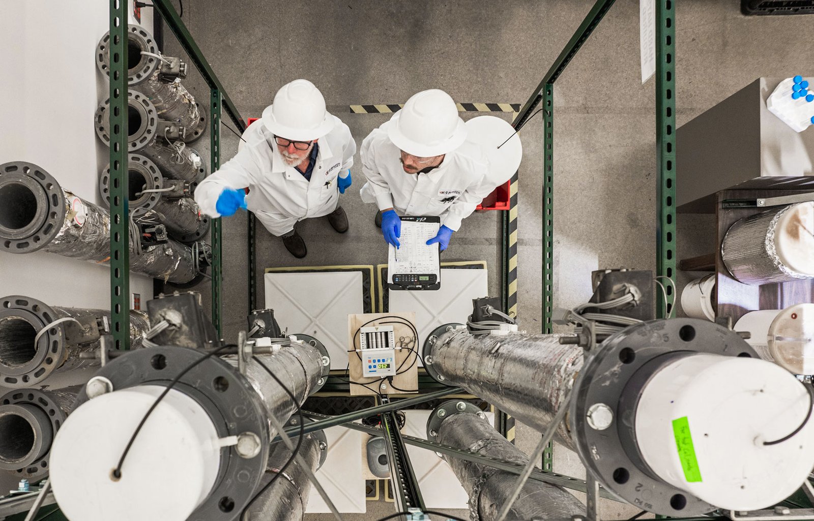 Two people in white coats and hard hats look up at steel columns inside a warehouse.
